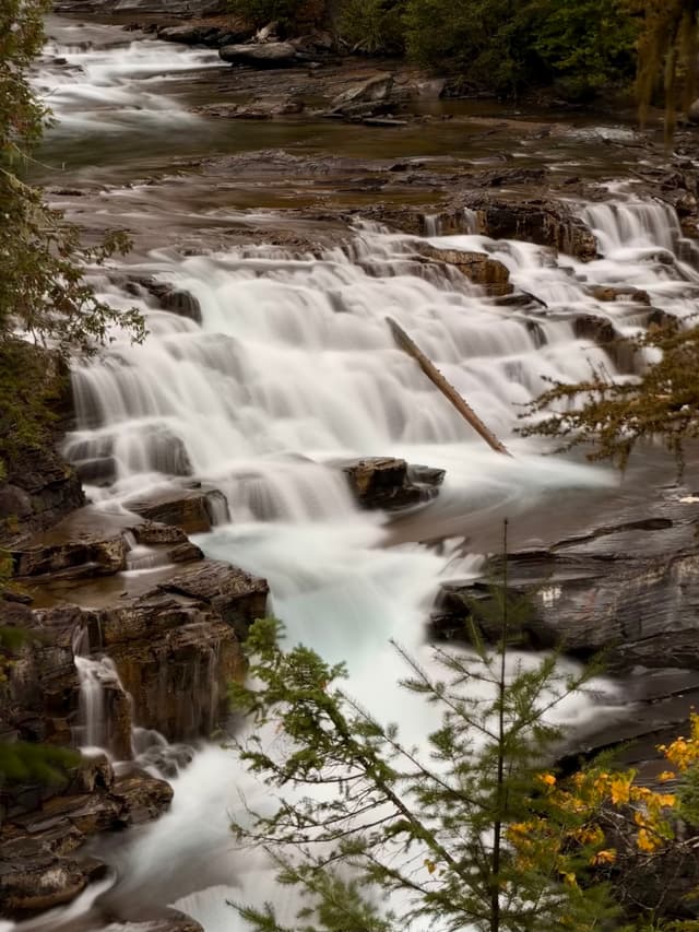 Multi-tiered waterfall cascading over rocky steps in a forest, captured with a silky long exposure; a fallen log crosses part of the flow and autumn foliage frames the scene