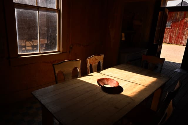A dimly lit room with a wooden table and chairs, featuring a bowl on the table. Sunlight streams through a window, casting shadows across the table