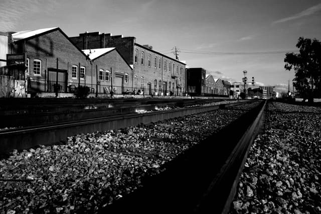 A black and white photograph of a railway track running alongside industrial buildings, with gravel covering the ground and a clear sky above