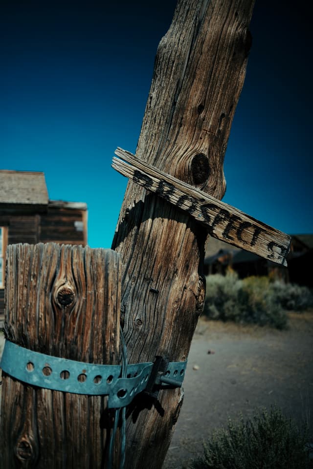 A weathered wooden post with a metal band and a broken sign, set against a clear blue sky and rustic buildings in the background