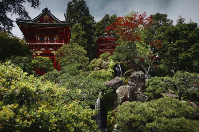 A traditional Japanese garden with lush greenery, a small waterfall, and a red pagoda-style building in the background