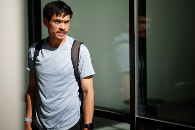 A man wearing a light blue shirt and backpack stands near a glass wall, with his reflection visible