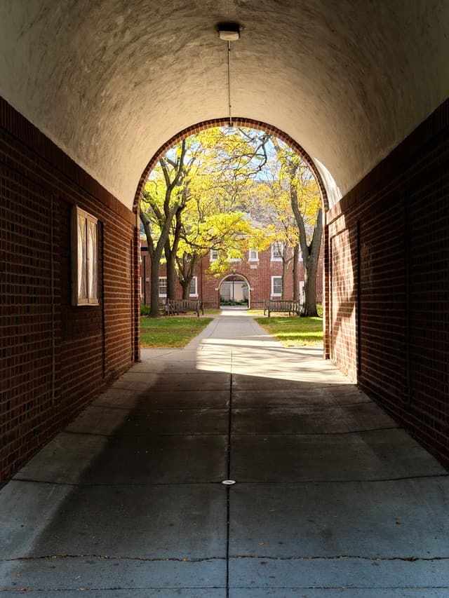 Brick archway framing a sunlit courtyard with trees and red-brick buildings