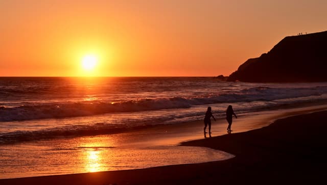 A sunset over the ocean with two people walking along the beach, silhouetted against the vibrant orange and red sky