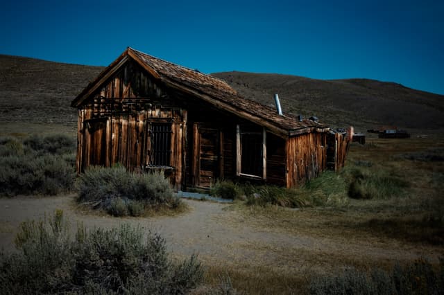 An old, weathered wooden cabin stands in a desolate, grassy landscape under a clear blue sky