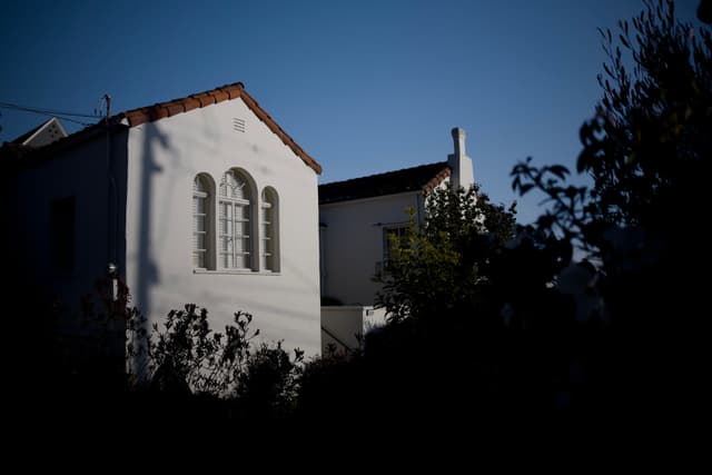 A white house with arched windows is partially illuminated by sunlight, surrounded by dark foliage and set against a clear blue sky