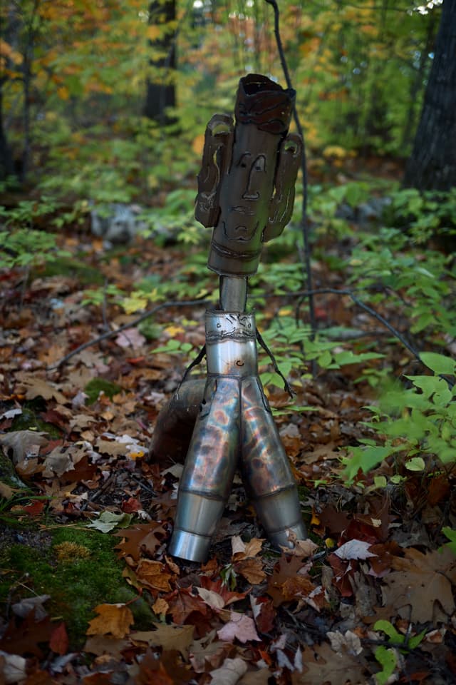 A small sculpture made from welded metal exhaust pipes, topped with a worn glove, standing among fallen leaves in a forest