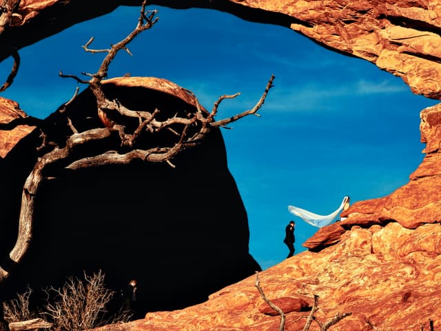 Bride and groom beneath a sandstone arch, veil flowing against deep blue sky