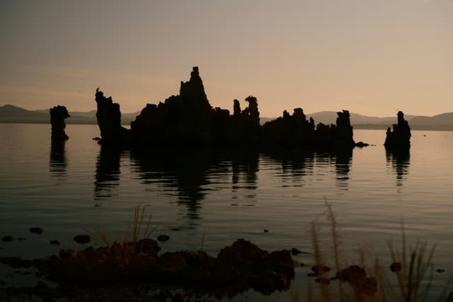 Silhouetted rock formations rise from a calm body of water at dusk, with distant mountains and a soft, dimly lit sky in the background