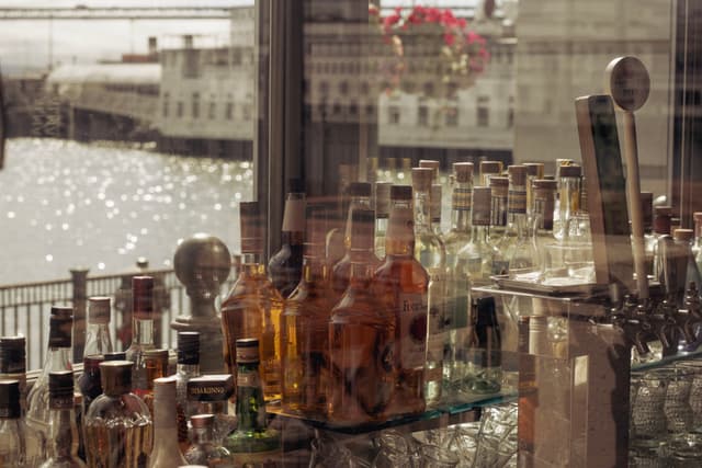 A collection of liquor bottles and bar taps is reflected in a window, with a waterfront view in the background