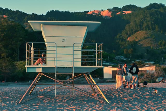 A lifeguard tower on a sandy beach with a person sitting inside. Nearby, a group of people is gathered around a small fire. The background features hills and trees under a clear sky