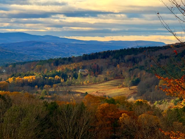 Sunlit valley with fall foliage on rolling hills, distant blue mountains, and a cloudy sky