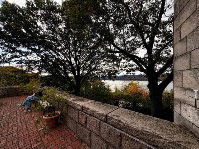 Brick patio beside stone wall with potted plant and a seated person, trees framing a river view