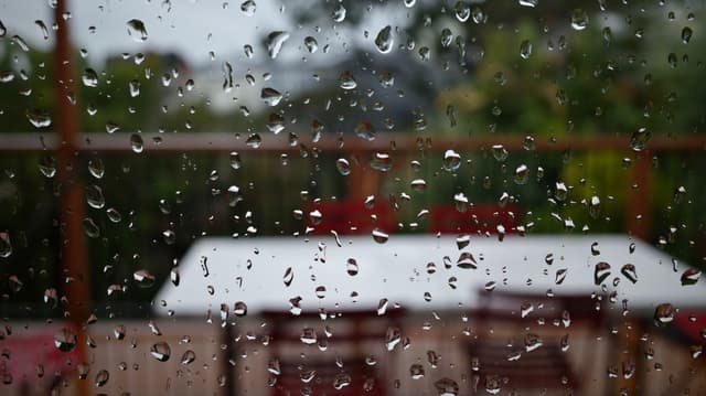 Raindrops on a window with a blurred view of outdoor furniture and greenery in the background