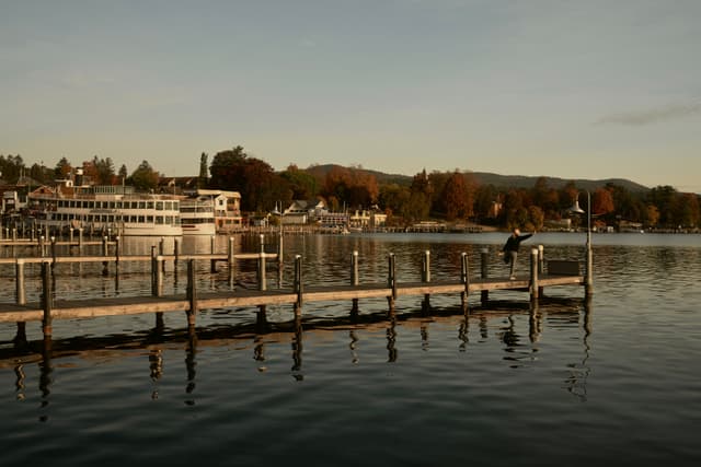 Wooden pier extending into a calm lake at sunset, with a moored ferry and lakeside buildings backed by autumn-colored hills