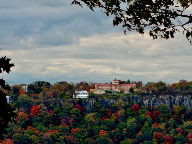 Large resort perched on a cliff above colorful autumn forest, framed by overhanging branches under a cloudy sky