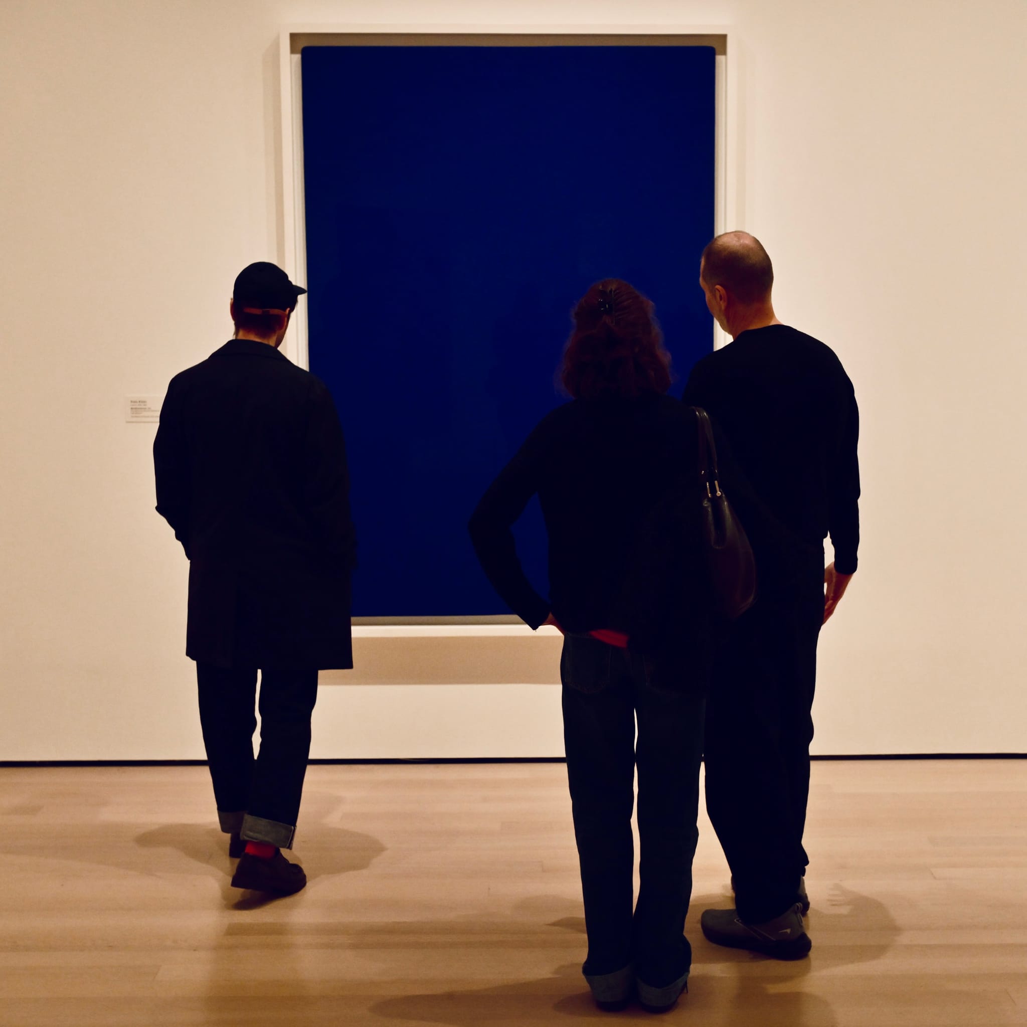 Three people in a gallery viewing a large dark blue painting on a white wall