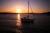 A sailboat on calm water during a vibrant sunset, with the sun low on the horizon and a silhouette of distant hills