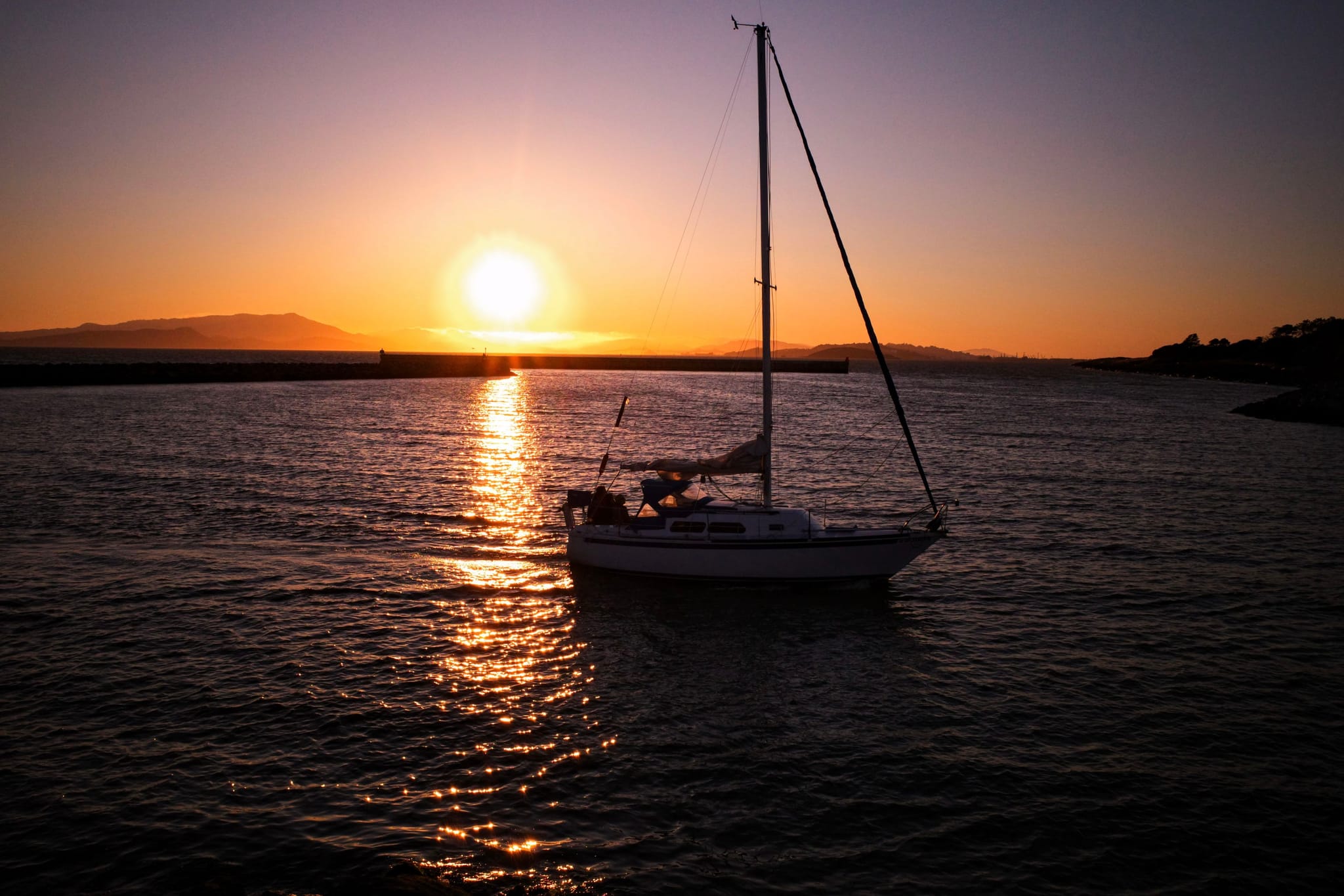 A sailboat on calm water during a vibrant sunset, with the sun low on the horizon and a silhouette of distant hills