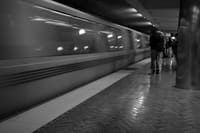 A black and white scene of a subway station with a train in motion and a few people standing on the platform