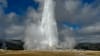 A powerful geyser erupts, sending a tall plume of water and steam into a cloudy sky over a rocky basin