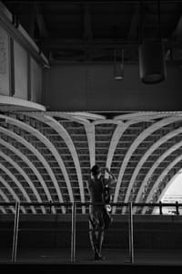 A person standing on a platform, silhouetted against an architectural structure with curved beams, in black and white