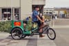 A man wearing a hat and a blue shirt rides a green pedicab on a city street