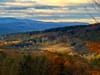Sunlit valley with fall foliage on rolling hills, distant blue mountains, and a cloudy sky