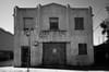 A black and white photo of an old, weathered two-story building with barred windows and a faded sign. The structure has a large central door and a smaller entrance on the side. Shadows and power lines are visible, adding to the urban atmosphere