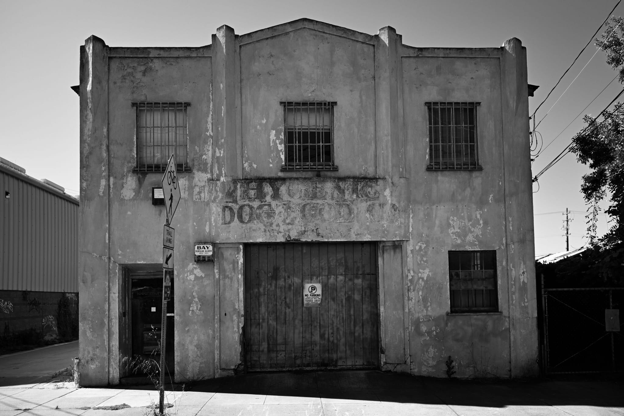 A black and white photo of an old, weathered two-story building with barred windows and a faded sign. The structure has a large central door and a smaller entrance on the side. Shadows and power lines are visible, adding to the urban atmosphere