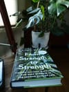 Book titled From Strength to Strength rests on a wooden desk beside a potted plant by a window