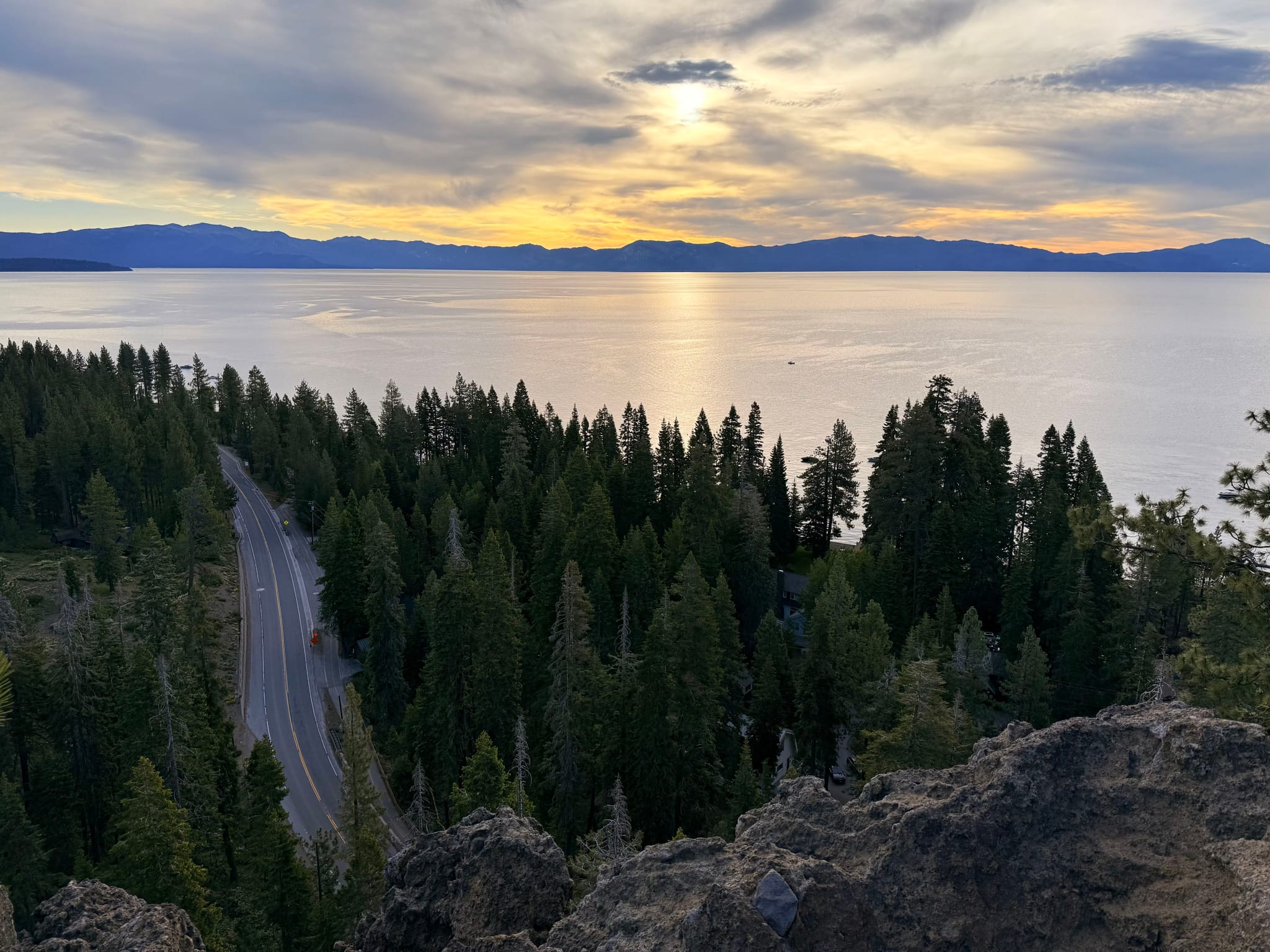 A scenic view of a lake at sunset, with a road winding through a forest of tall trees and mountains in the background