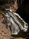 A tiered waterfall cascading over dark rock in a narrow canyon, captured with a long exposure, with pine trees along the rim