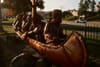 Wooden sculpture of several figures paddling a canoe, set outdoors near a roadside fence in warm late-day light