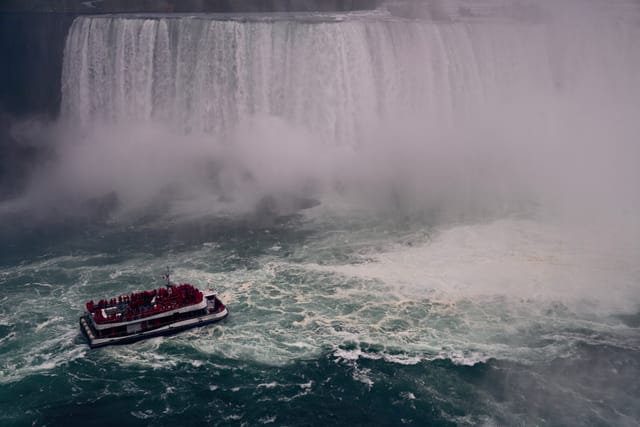 A red-and-white tour boat approaches the misty base of a massive waterfall, likely Niagara Falls