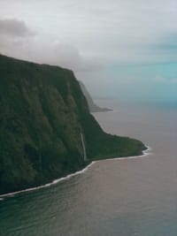 Steep green cliffside along the coastline with a waterfall cascading into the sea and a distant horizon
