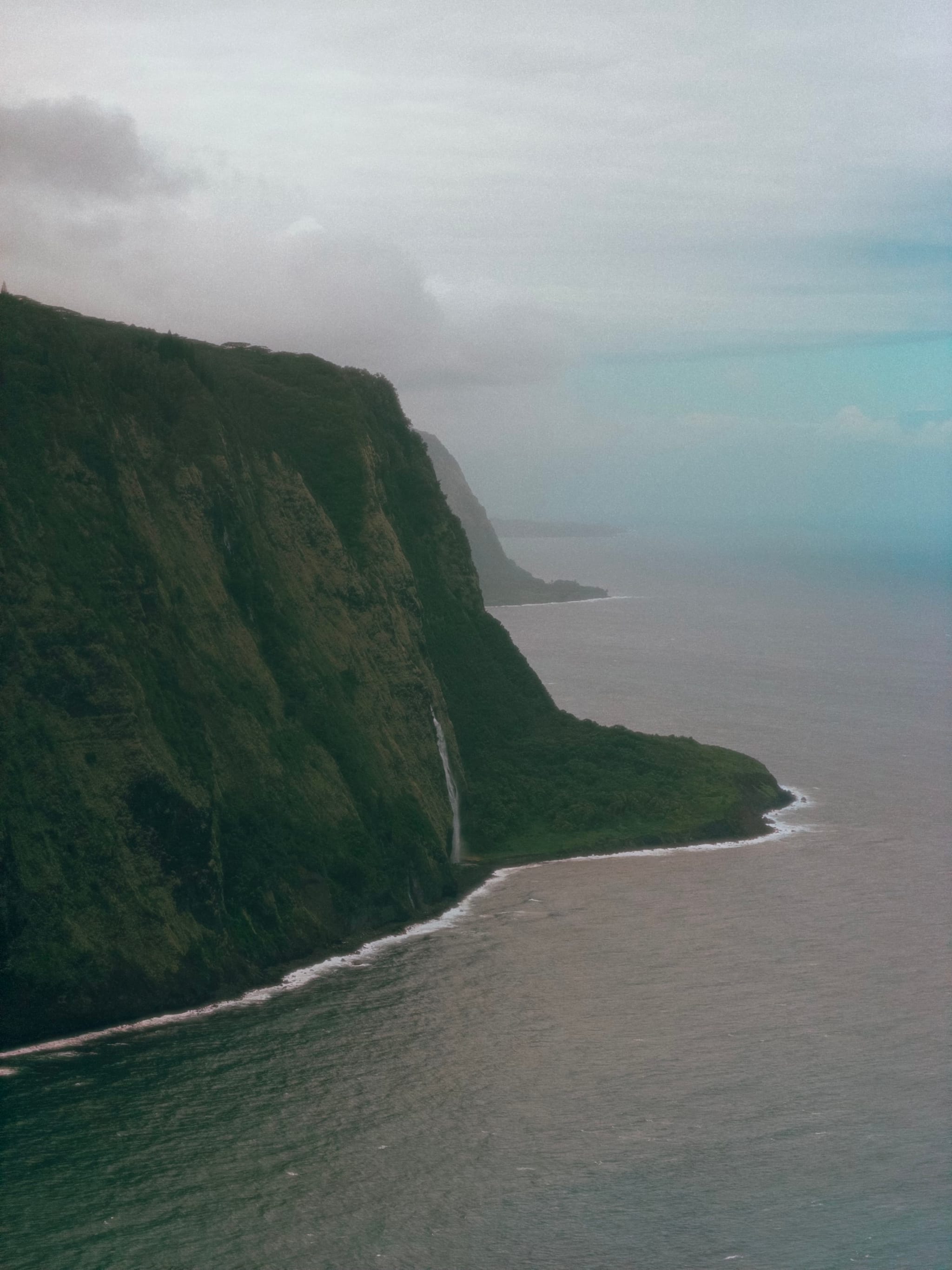 Steep green cliffside along the coastline with a waterfall cascading into the sea and a distant horizon