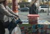 A street vendor pushes a cart covered with images of ice cream, carrying a red-lidded container and a bag, while a person walks by on the sidewalk
