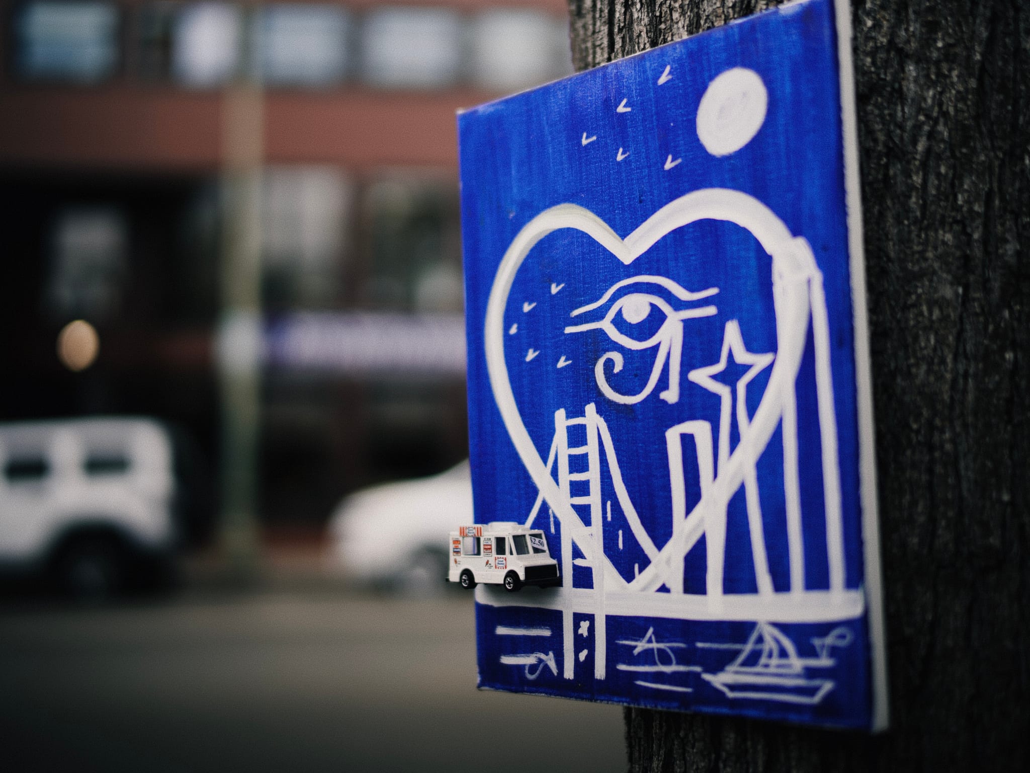 A blue and white artwork attached to a tree, featuring a heart with an eye, a bridge, and cityscape elements, with a small toy vehicle on the side