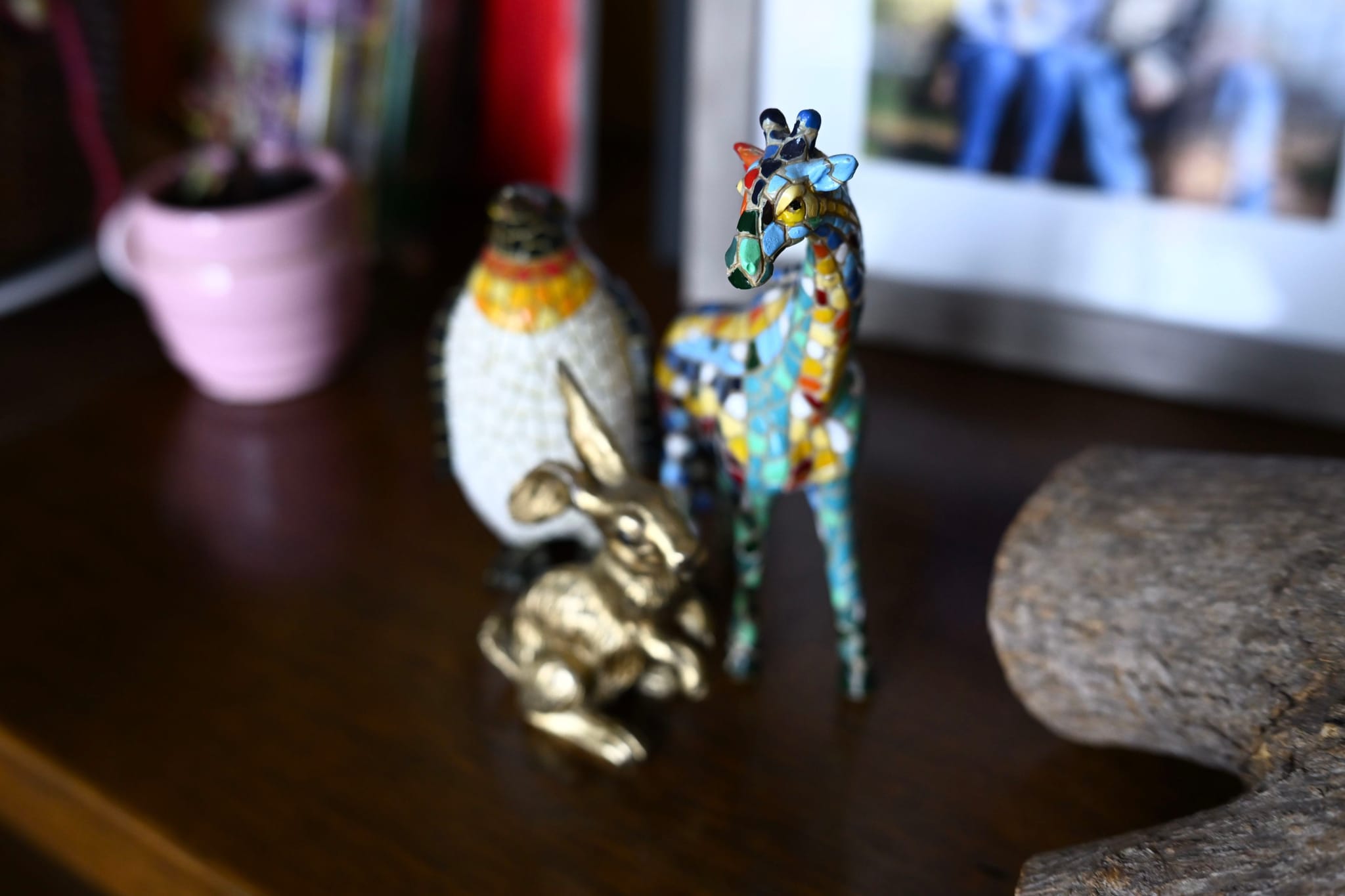 A small collection of decorative items on a wooden surface, including a colorful giraffe figurine, a golden rabbit, and a textured vase, with a framed photo in the background