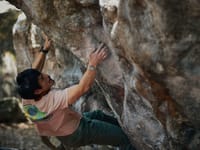A person bouldering on a large rock formation, wearing a light-colored shirt and green pants, focused on climbing