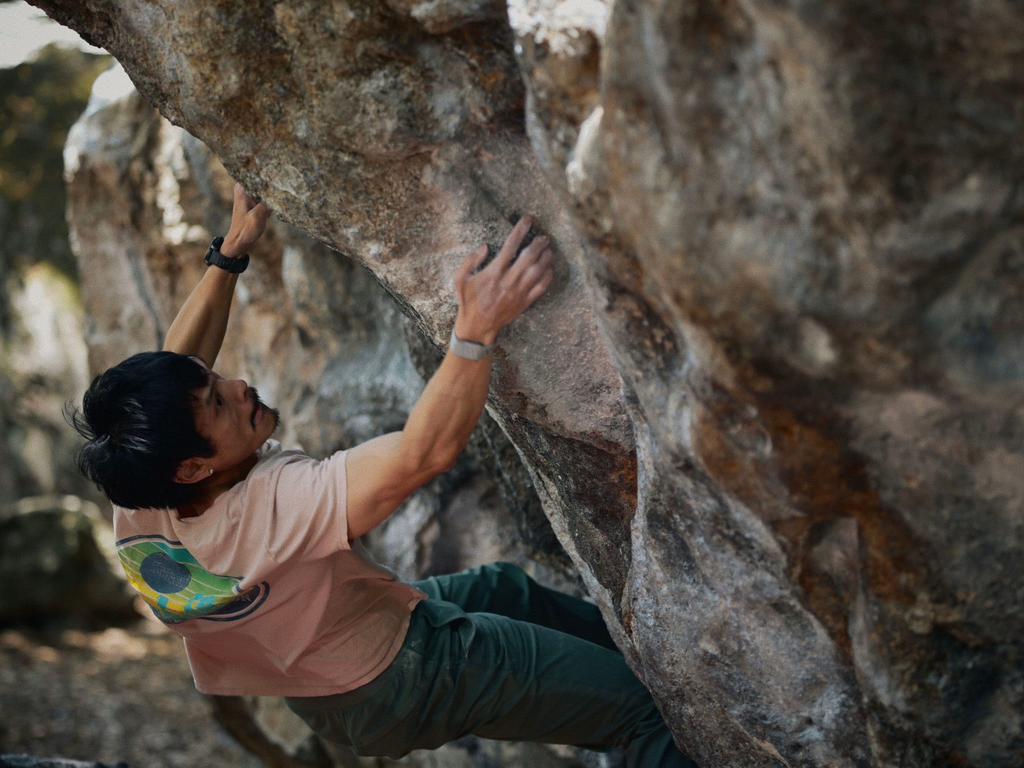 A person bouldering on a large rock formation, wearing a light-colored shirt and green pants, focused on climbing