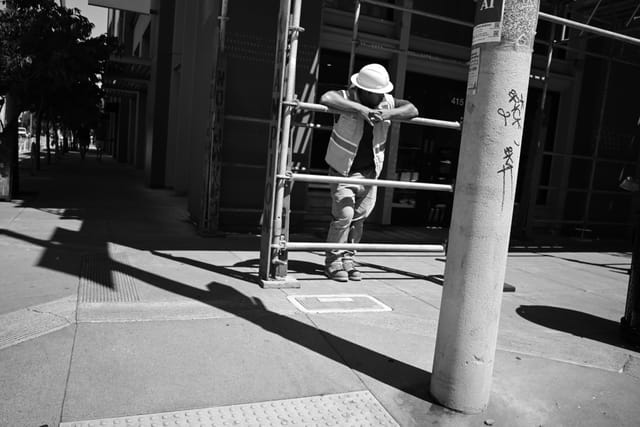 A construction worker in a hard hat and safety vest leans on scaffolding at a street corner, with strong shadows cast on the sidewalk. The scene is in black and white