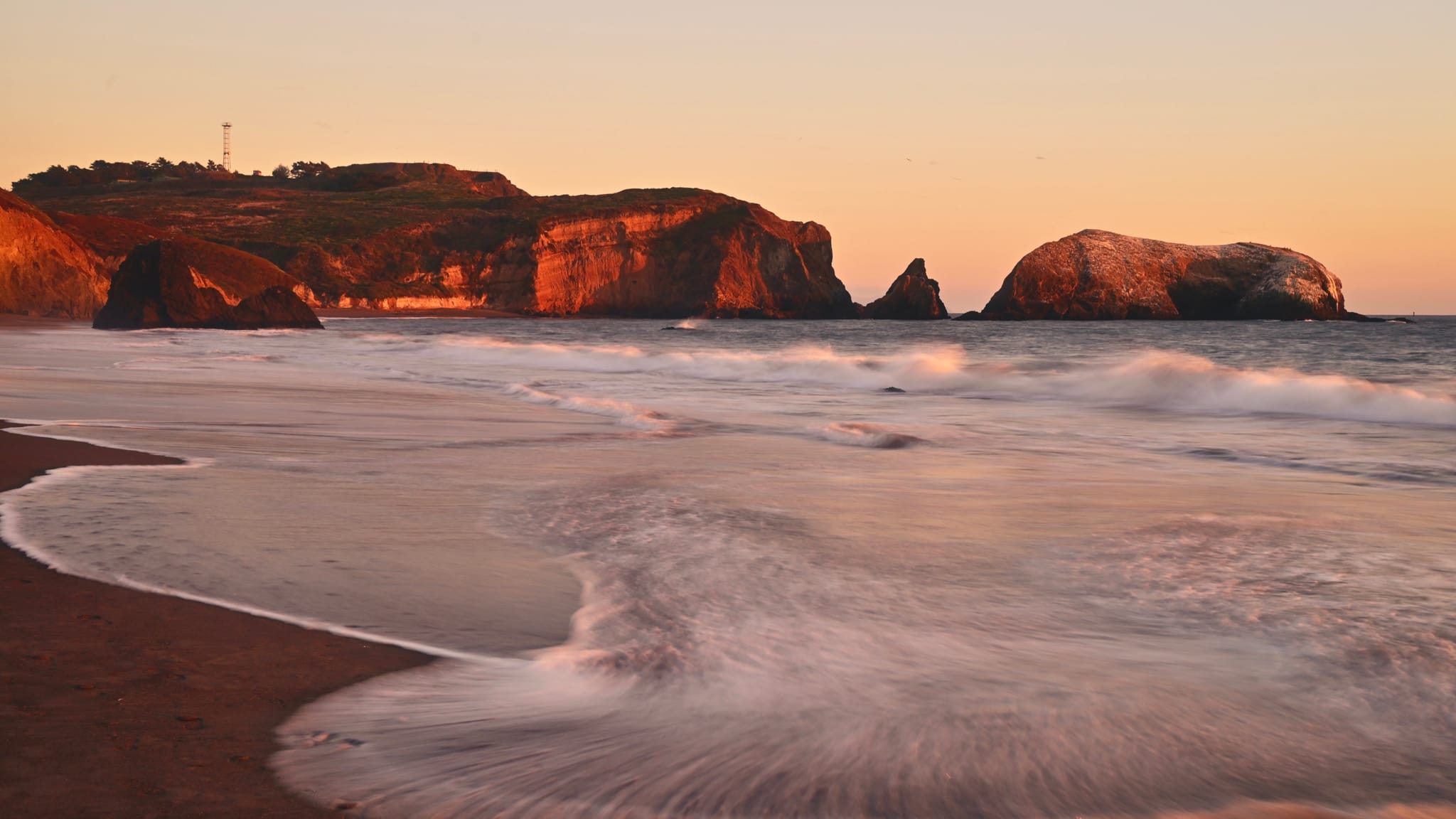 A serene coastal scene at sunset with gentle waves washing onto a sandy beach, rocky cliffs in the background, and a warm, golden glow illuminating the landscape