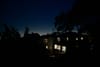 A nighttime scene with houses illuminated from within, surrounded by silhouetted trees against a dark blue sky