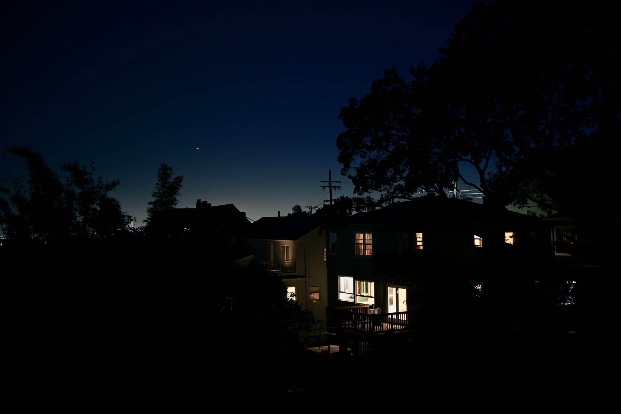 A nighttime scene with houses illuminated from within, surrounded by silhouetted trees against a dark blue sky