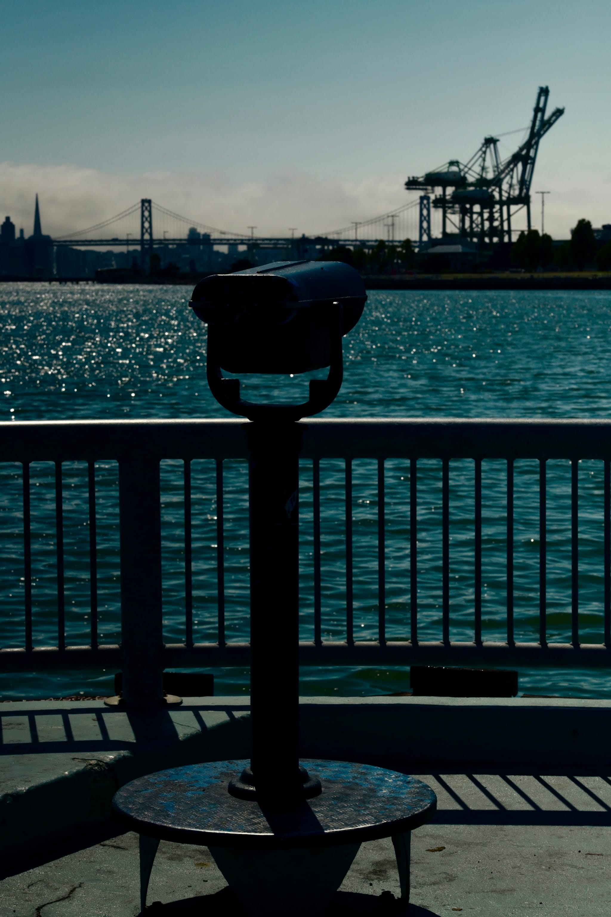 A waterfront scene with a coin-operated binocular viewer in the foreground, a body of water, a bridge, and cranes in the background