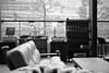 A black and white interior scene with a leather sofa, a wooden table, and a shelf with various items. Large windows reveal an urban view with trees and buildings outside
