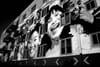 A black and white mural on a building features large, expressive faces of smiling individuals, with windows integrated into the artwork