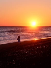 A person walking along a beach at sunset, with the sun low on the horizon and the sky filled with warm orange hues